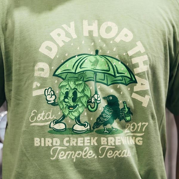 Close-up of a green Bird Creek Brewing t-shirt with a cartoon hop holding an umbrella.