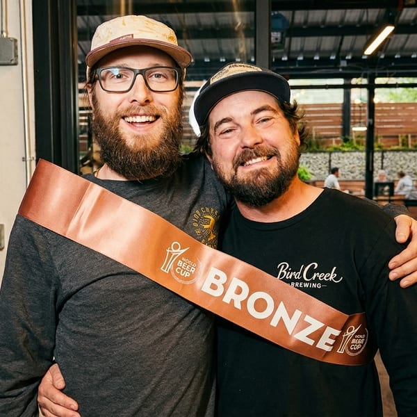 Two smiling men, one wearing a "World Beer Cup Bronze" sash, embracing in a brewery setting.