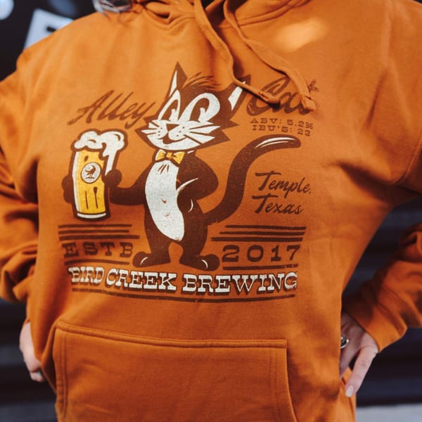 Close-up of an orange hoodie featuring a cartoon cat holding a beer mug, with "Alley Cat" and "Bird Creek Brewing" text.